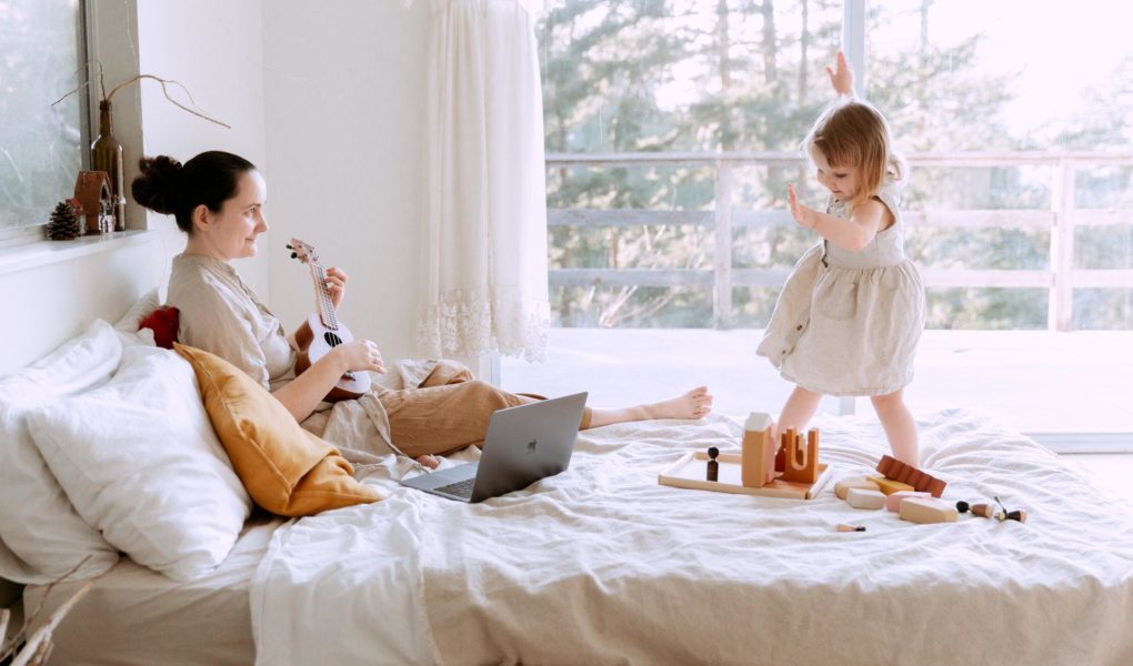 A mother with a toddler on a bed playing the ukulele to keep her entertained.