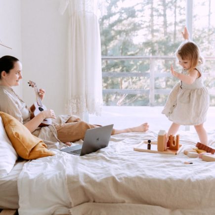 A mother with a toddler on a bed playing the ukulele to keep her entertained.