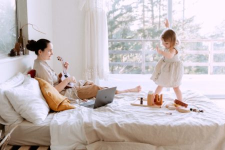 A mother with a toddler on a bed playing the ukulele to keep her entertained.